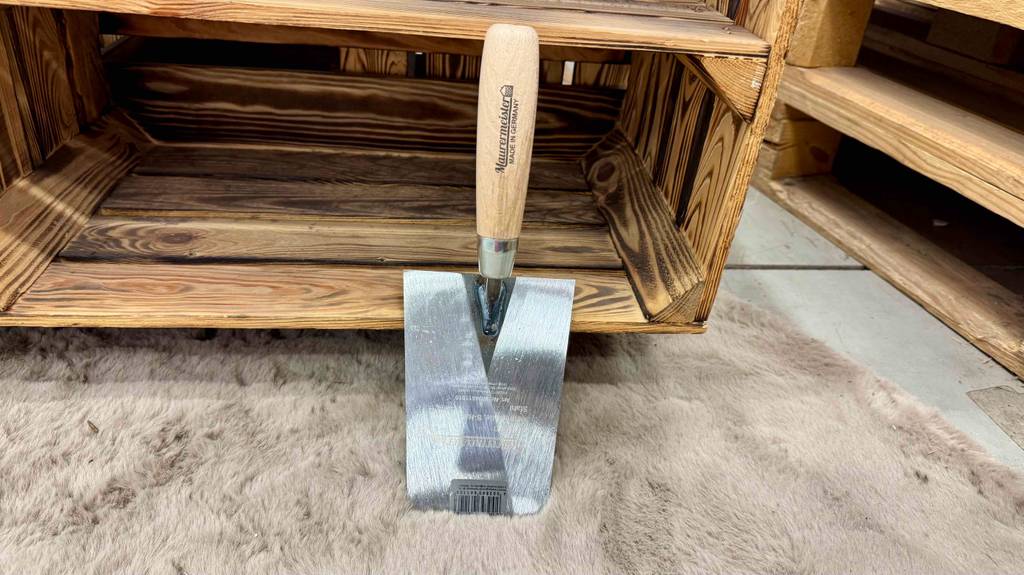 Die getestete Maurerkelle mit Holzgriff lehnt an einem Holzregal.