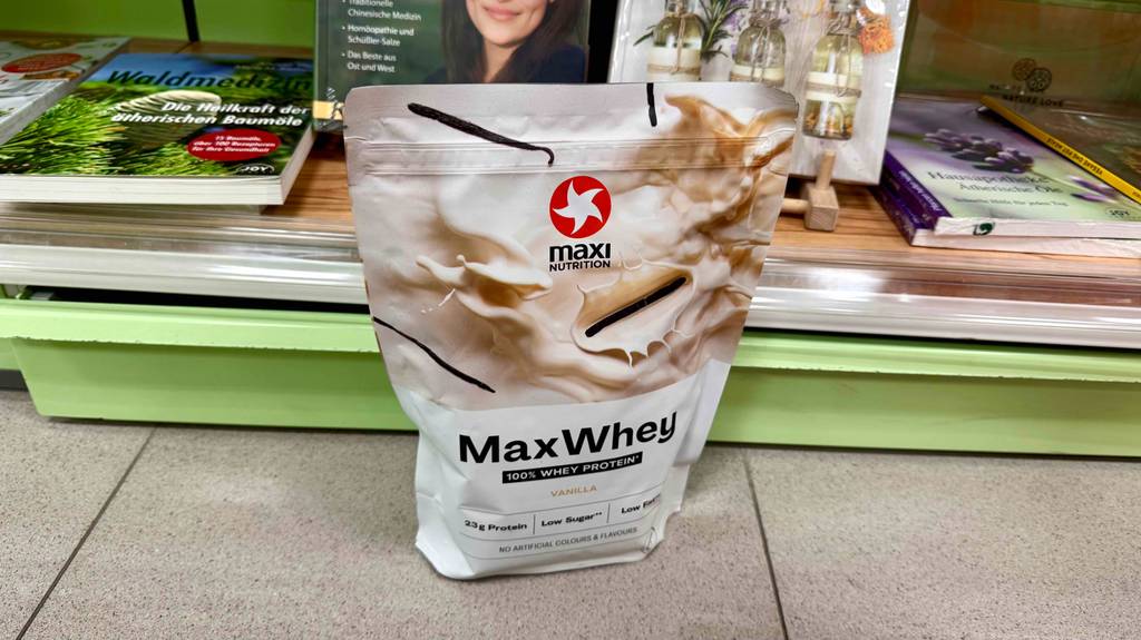 Eine Packung Whey-Protein-Vanille steht zum testen vor einem Bücherregal.