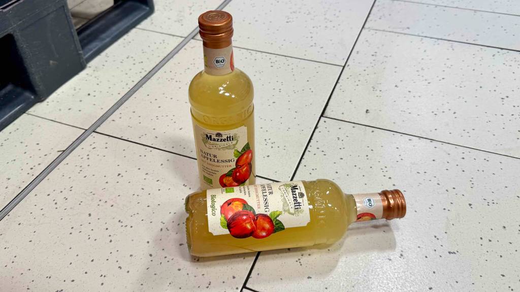 Eine Flasche getesteter Apfelessig naturtrüb liegt quer vor einer stehenden auf einem Fliesenboden.