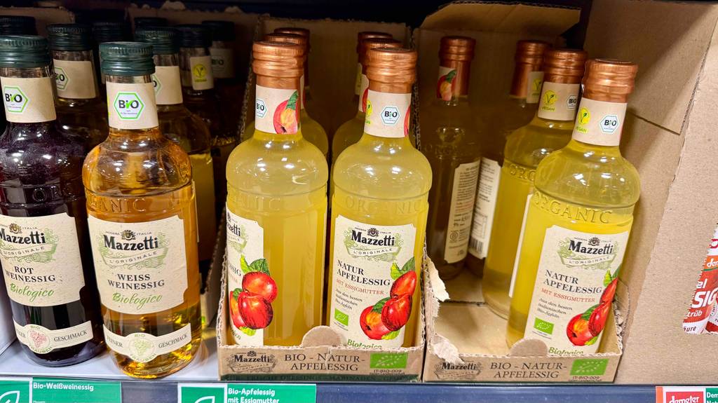 Mehrere Flaschen Apfelessig naturtrüb stehen zum Testen neben- und hintereinander in einem Regal neben anderen Produkten.