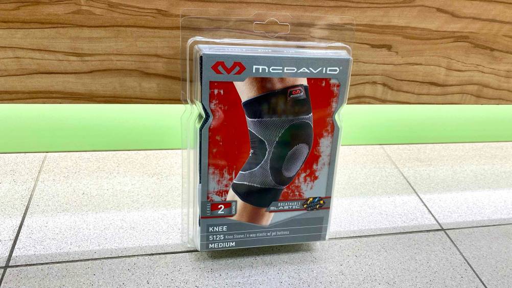 Kniebandage getestet: Eine originalverpackte Bandage steht auf einem Fließenboden, im Hintergrund eine grüne Fläche und eine Holzwand.