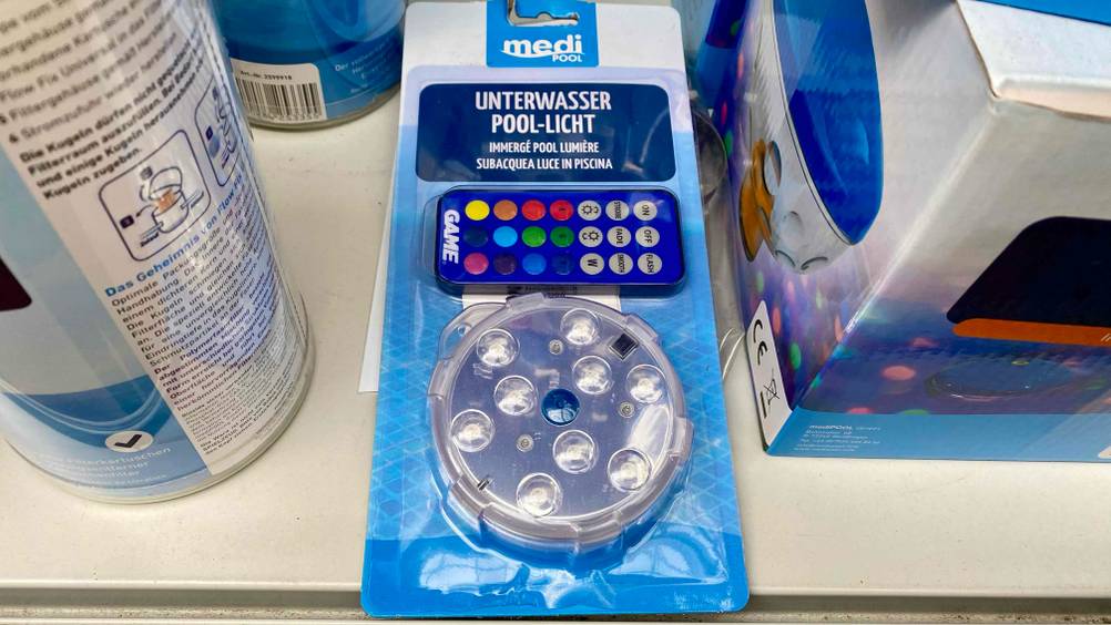 Eine getestete Poolbeleuchtung in Originalverpackung inklusive Fernbedienung liegt zwischen anderen Produkten auf einem hellen Regal.