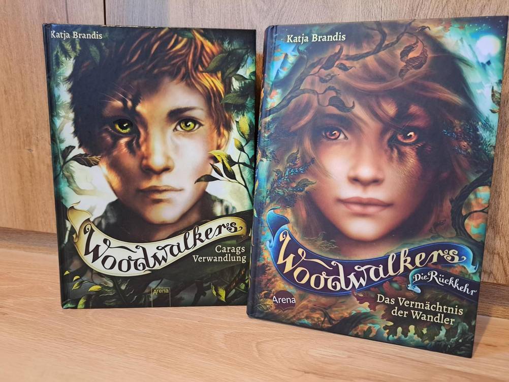 Zwei Bücher der Woodwalkers-Romanreihe stehen neben einander. Auf dem linken Roman ist die Titelfigur Carag zu sehen, dessen rechts Gesichtshälfte teilverwandelt in einen Puma ist. Auf dem rechten Buch ist ebenfalls ein Junge zu sehen, dessen linke Gesichtshälfte teilverwandelt in eine Raubkatze ist.