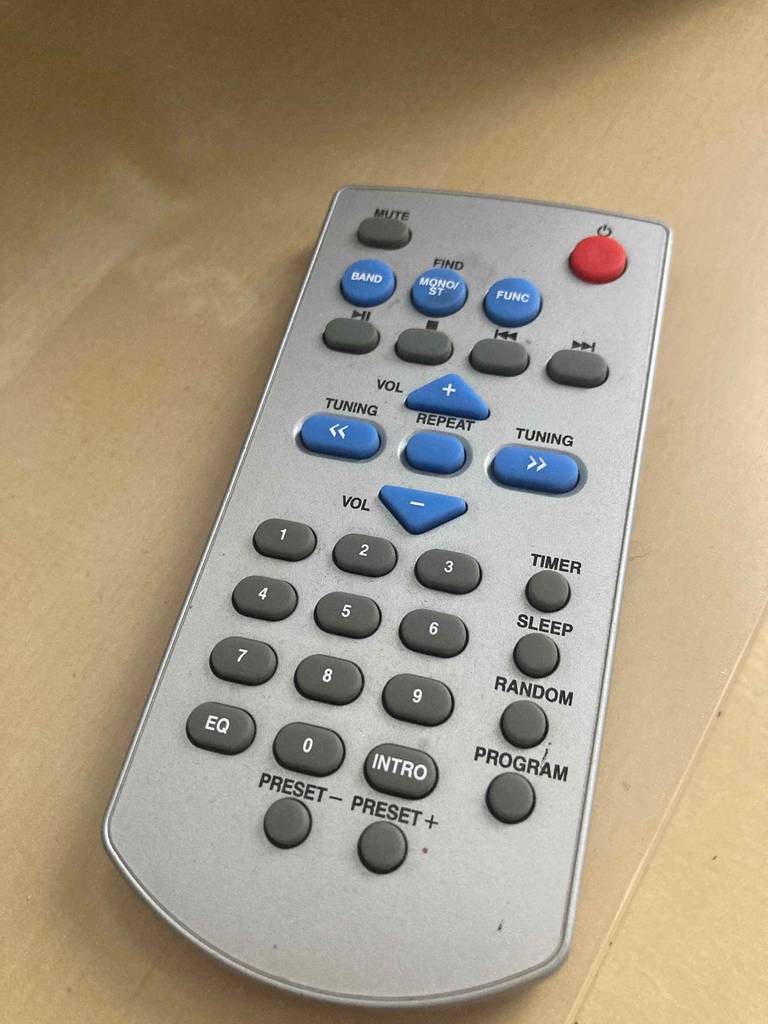 Hellgraue Fernbedienung von einem CD-Player mit Kassettendeck im Test liegt auf beigefarbigem Untergrund.