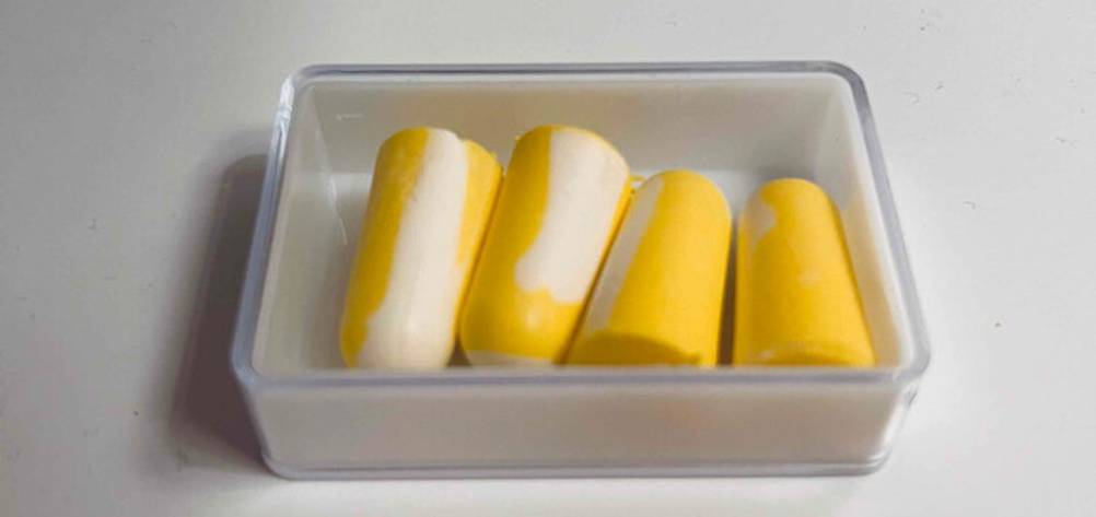 Vier weiche, gelb-weiße getestete Ohrstöpsel liegen in einer offenen Aufbewahrungsbox.