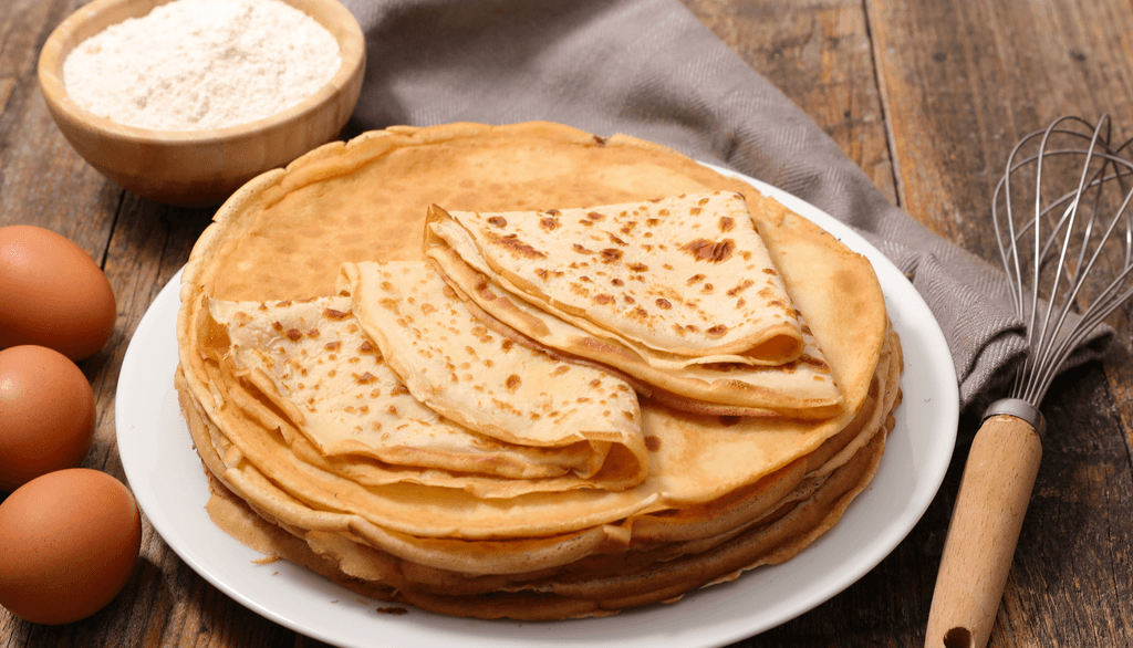 Mehl-00-Test: Crêpes auf einem Teller, daneben befindet ich eine Schale Mehl, Eier und ein Besen. 