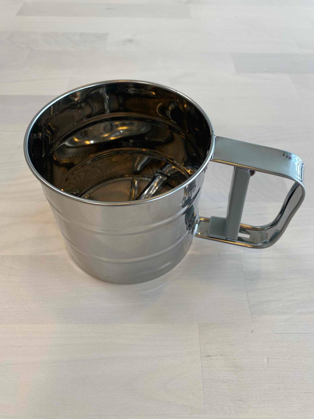 Ein Mehlsieb mit Henkel stehend auf einer hölzernen Fläche.