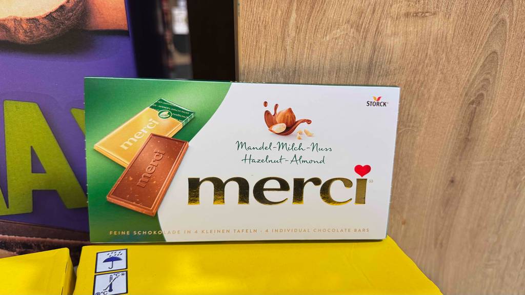 Eine Tafel der Sorte Mandel-Milch-Nuss steht auf einem Karton angelehnt an ein Holzregal.