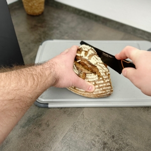 Ein Brotmesser aus dem Fiskars-6424009787917-Set wird verwendet, um ein rundes Brot auf einem Schneidebrett zu schneiden. Beide Hände führen das Messer präzise.