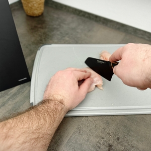 Ein Fleischstück wird mit einem Messer aus dem Fiskars-6424009787917-Set auf einem Schneidebrett zerteilt. Der Messerblock steht im Hintergrund.