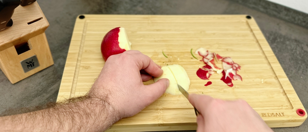 Ein Apfel wird mit einem Messer aus dem WMF-1874699990-Set auf einem Holzbrett geschnitten. Der Messerblock steht im Hintergrund.