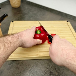 Eine rote Paprika wird mit einem Messer aus dem WMF-1874699990-Set auf einem Holzbrett zerteilt. Der Schnitt erfolgt mittig durch das Gemüse.
