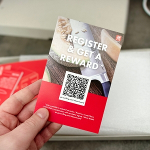 Ein Beileger aus dem Zwilling-Set mit Hinweis zur Registrierung und QR-Code wird in der Hand gehalten. Verpackung ist im Hintergrund sichtbar.