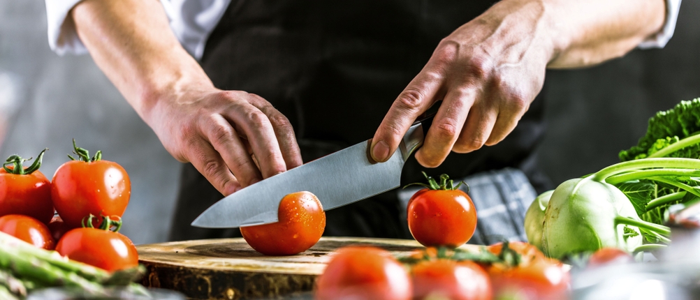 Messerschärfer-Test: Eine Person schneidet mit einem großen Messer Tomaten auf einem Holzbrett. Um das Brett herum sind Tomaten, Kohlrabi und weitere Lebensmittel aufgestellt.