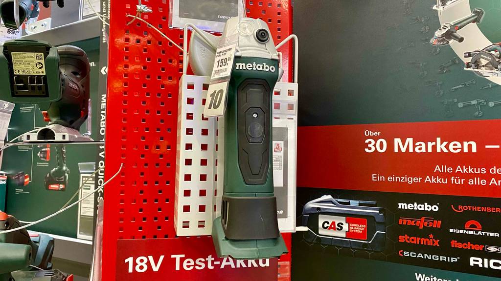 Winkelschleifer-Test: ein grün-schwarzer Metabo Winkelschleifer ohne Akku inklusive Schutzhaube aus Metall befindet sich an einem roten Ausstellungsregal in einem Metabo-Bereich innerhalb eines Geschäfts.
