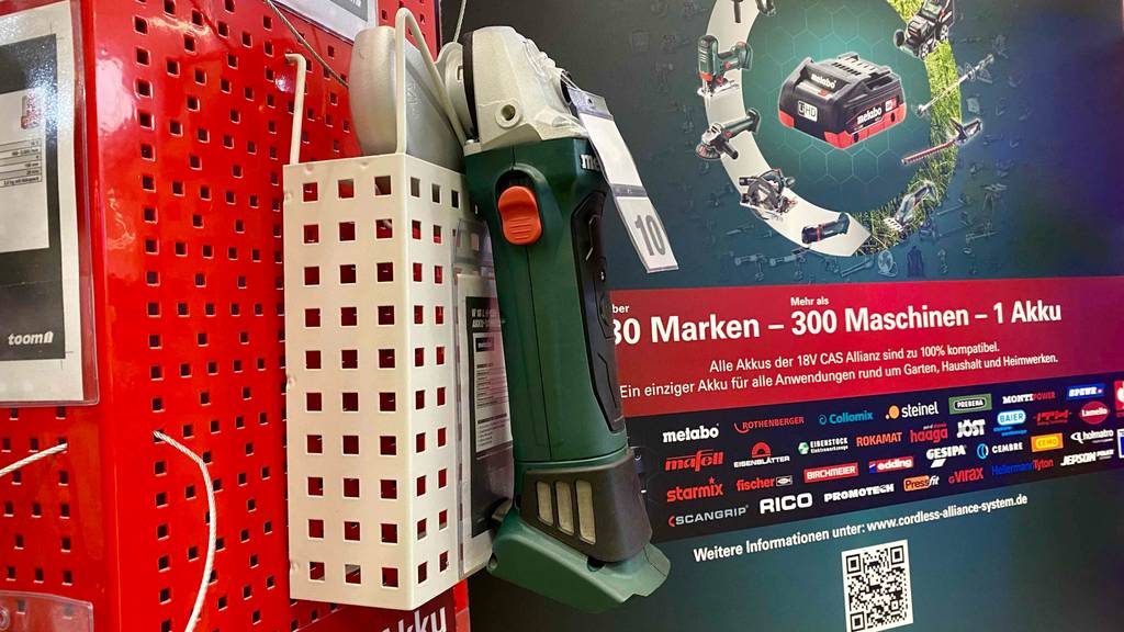 Winkelschleifer-Test: Seitenansicht auf einen grün-schwarzen Winkelschleifer ohne Akku an einem roten Ausstellungsregal, mit Schutzhaube aus silberfarbenem Metall und Metabo-Akku Werbeposter im Hintergrund.