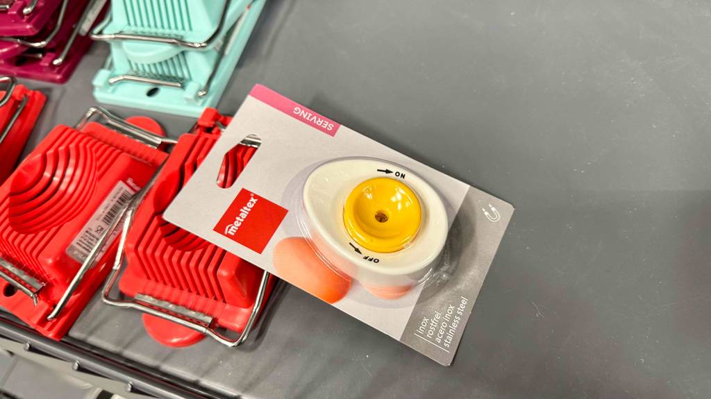 Im Test: Der Eierstecher in Weiß-Gelb in der Verpackung liegt auf einer grauen Fläche, auf der sich rote Eierschneider befinden.