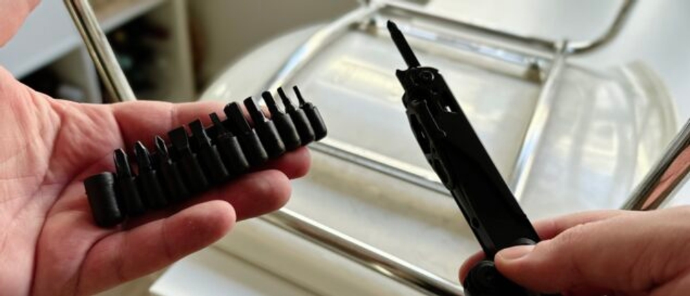 Eine Hand hält das Bitset des Miaoke 23-in-1 Multitool vor einem umgedrehten Metallstuhl. Das Multitool mit eingesetztem Bit ist ebenfalls zu sehen.