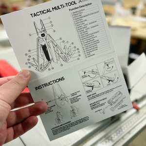 Eine Hand hält die Bedienungsanleitung des Miaoke 23-in-1 Multitool mit einer schematischen Darstellung der Funktionen. Der Hintergrund zeigt eine Werkstatt mit Werkzeugen.
