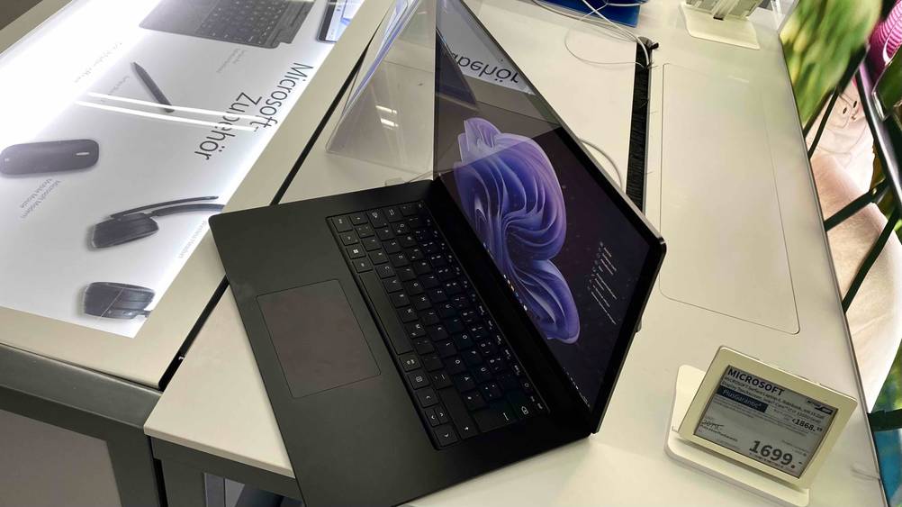 Seitenansicht von oben auf ein schwarzes geöffnetes sowie eingeschaltetes Microsoft Surface 15-Zoll-Laptop im Test inklusive Hochglanz-Display neben seinem Preisschild.