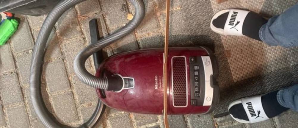 Füße in weißen Puma-Badelatschen neben einem weinroten Miele-Tierhaarstaubsauger im Test auf grauem Pflasterklinker.