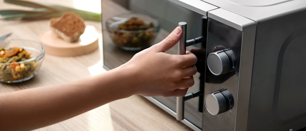 Mikrowelle ohne Drehteller im Test: Eine Hand öffnet eine Mikrowelle. Im Hintergrund stehen Brot und eine Schüssel mit Gemüse.