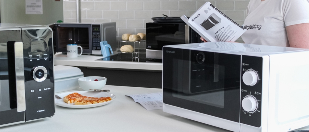 Mehrere Mikrowellen im Test bei Vergleich.org stehen auf einem Tisch neben einem Teller mit Pizza.