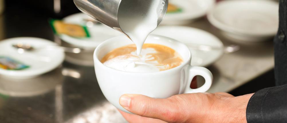 Milchaufschäumer-Bodum-Test: Barista gießt Milchschaum aus einer Kanne in ein Kaffee-Heißgetränk ein