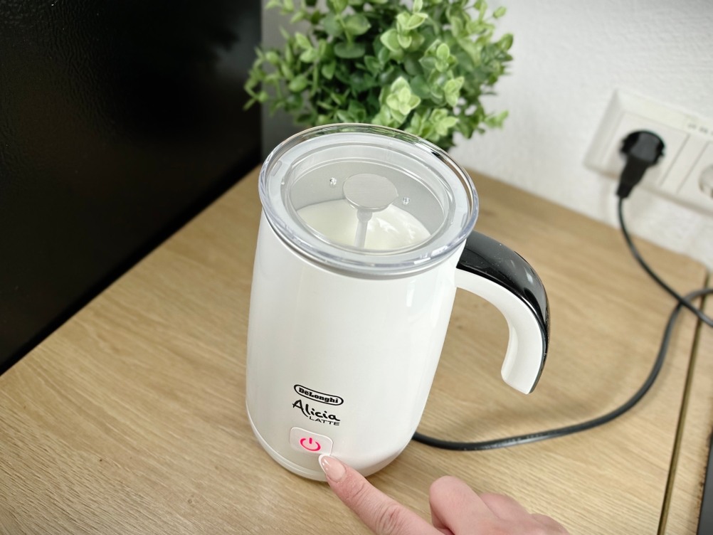 Ein Finger drückt den Einschaltknopf des De'Longhi EMF 2.W Alicia Latte Milchaufschäumers. Das Gerät steht auf einer Holzoberfläche in einer Küche.
