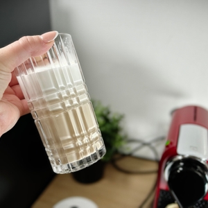 Ein Latte Macchiato mit Milchschaum wird in einem Glas gehalten. Im Hintergrund steht eine rote Nespresso-Kapselmaschine auf der Arbeitsplatte.