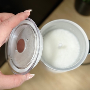 Eine Hand hebt den Deckel vom Milchaufschäumer De'Longhi EMF 2.W an. Im Inneren ist frisch aufgeschäumte Milch zu sehen.
