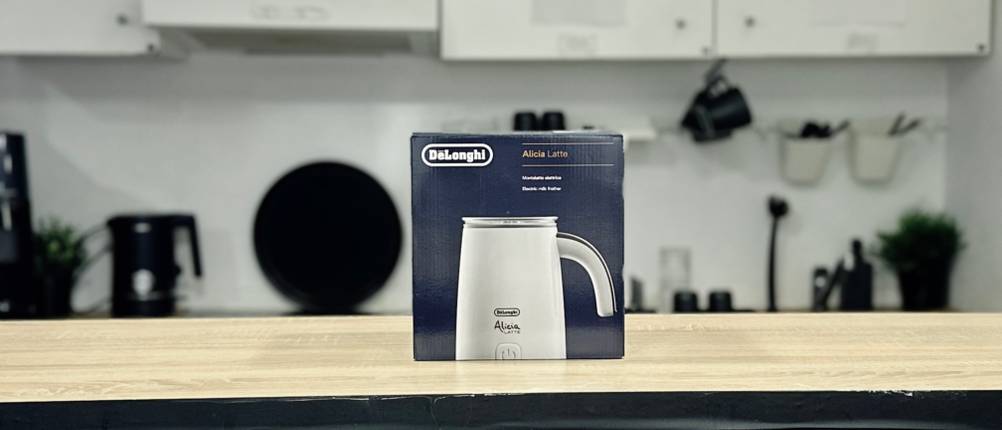 Die Verpackung des De’Longhi EMF 2.W Alicia Latte steht auf einem Holztresen. Im Hintergrund ist eine moderne Küche mit weißen Schränken zu sehen.