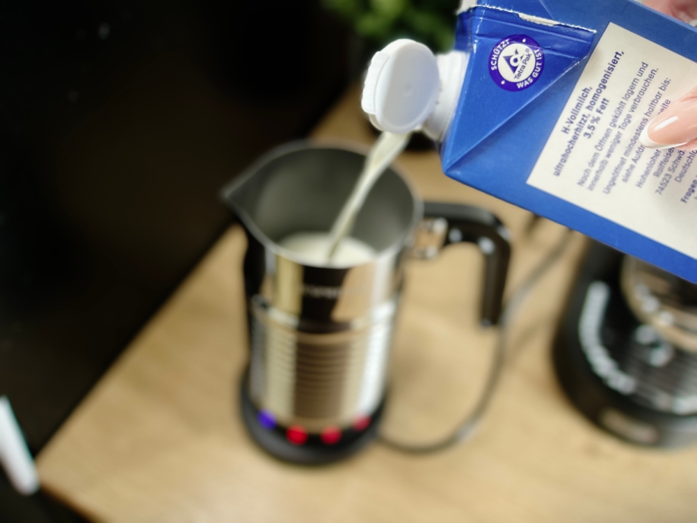 Milch wird aus einem blauen Karton in den Nespresso Aeroccino 4 gegossen. Das Gerät steht offen auf einer Küchenplatte.