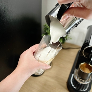 Der Milchschaum wird aus dem Nespresso Aeroccino 4 in ein Glas gefüllt. Die Kaffeemaschine steht im Hintergrund.
