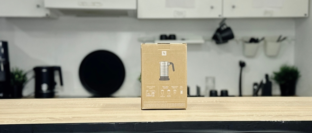 Die Verpackung des Nespresso Aeroccino 4 steht auf einer Holztheke. Im Hintergrund ist eine Küchenzeile mit Hängeschränken sichtbar.