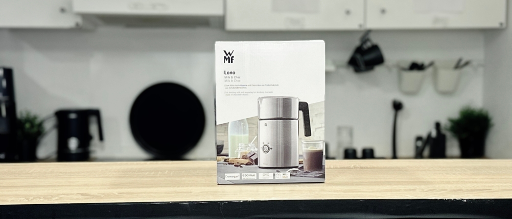 Die Verpackung des WMF Lono Milk & Choc steht auf einem Holztresen vor einer Küchenzeile. Die Box zeigt Produktbild und Anwendung.