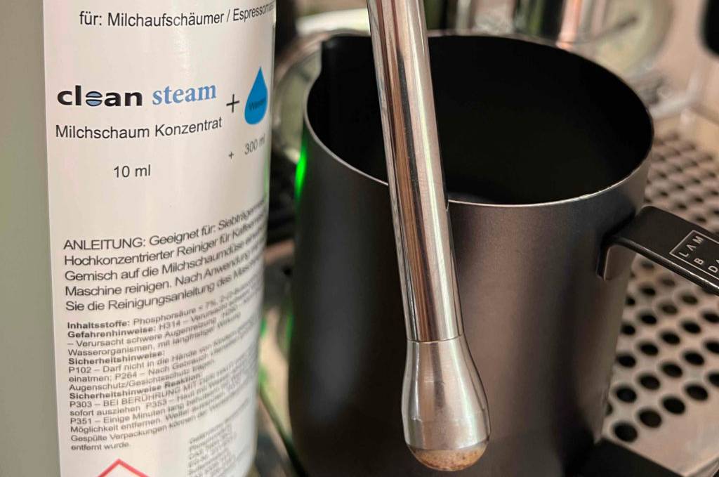 Eine Flasche getesteter Milchsystemreiniger wird neben eine Aufschäumdüse und ein Kännchen gehalten.