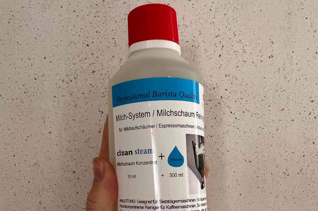 Milchsystemreiniger getestet: Eine Hand hält eine Flasche vor eine helle Wand.