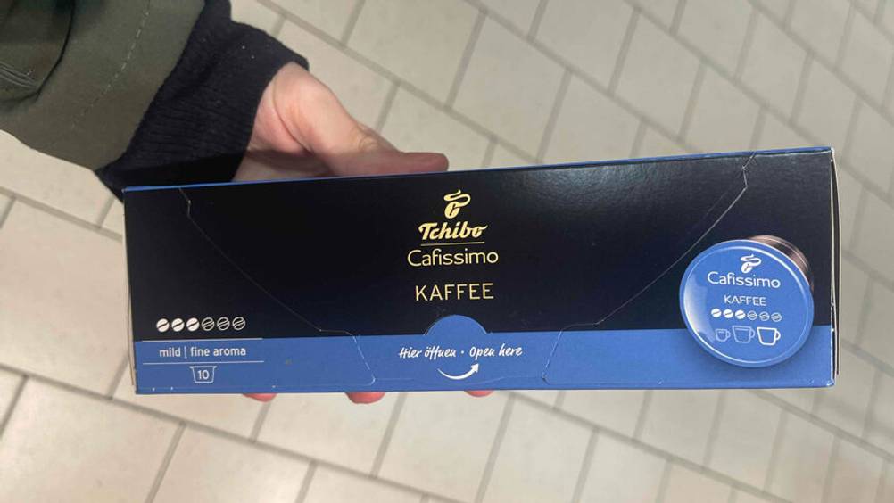 Eine Hand hält die schwarz-blaue Packung inklusive zehn der getesteten Cafissimo-Kapseln „Mild“ über einem weißen Fliesenboden.