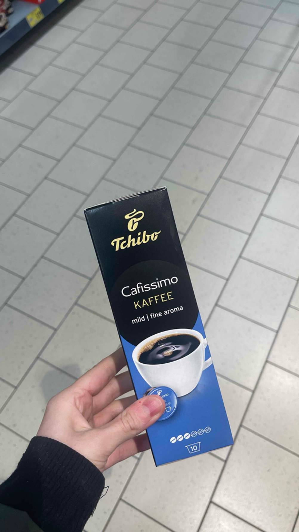 Eine Hand hält die schwarz-blaue Packung inklusive der Cafissimo-Kapseln im Test über einem weißen Fliesenboden.