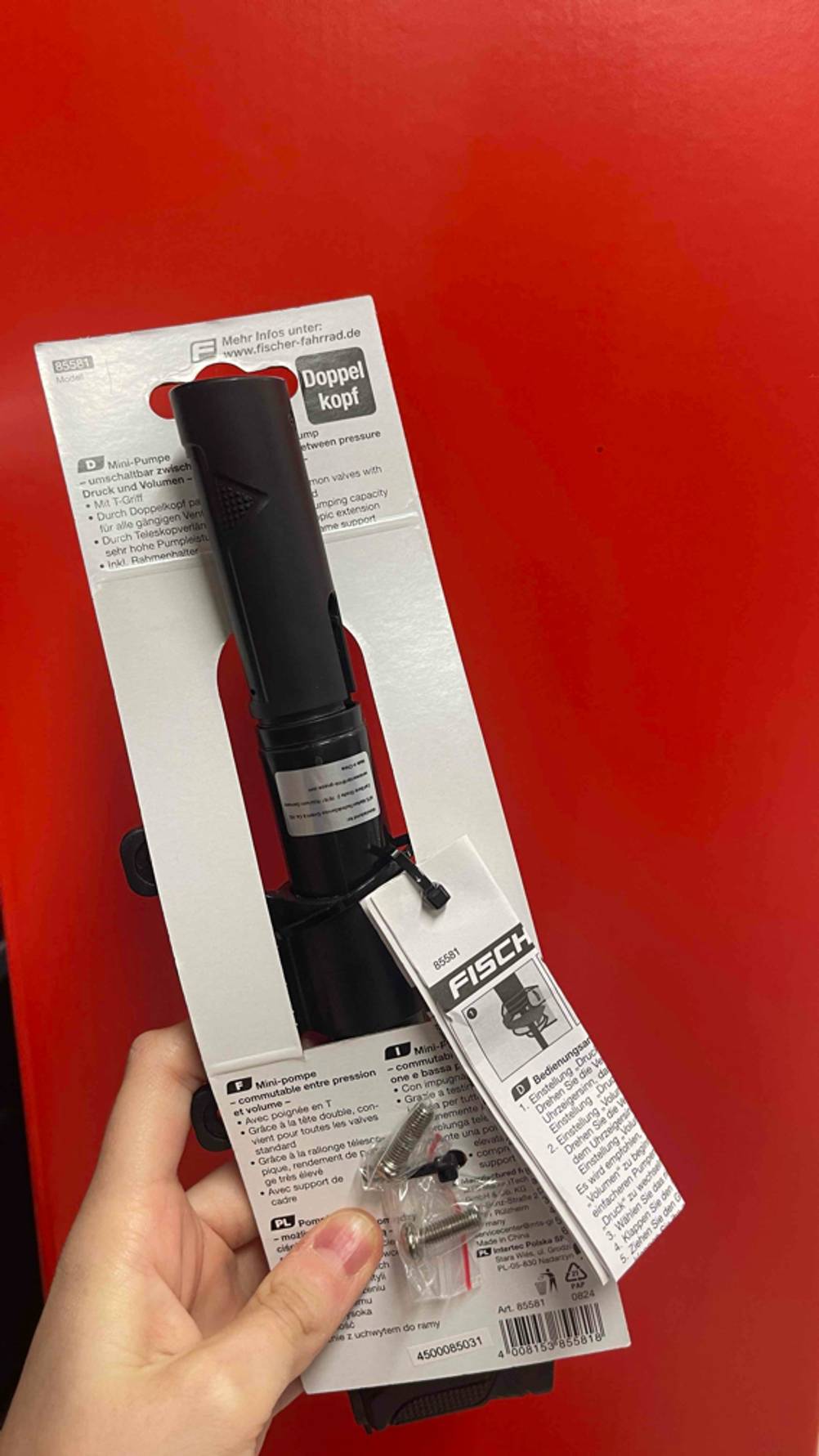Eine Hand hält die Rückseite der verpackten Minipumpe im Test vor eine rote Wand.