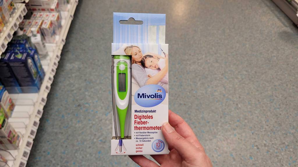 Ein digitales Fieberthermometer im Test in einer Hand über dem Boden eines Drogeriemarktes.