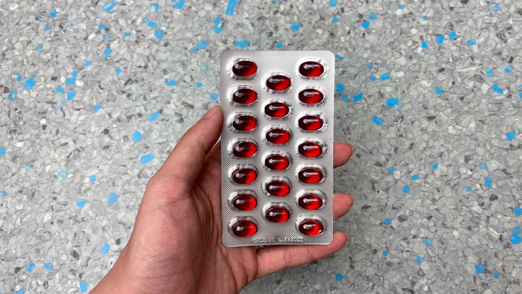 Getestet. Hand hält einen Blister mit 20 Kapseln Krillöl vor grau-blau-schwarz-gemustertem Hintergrund.