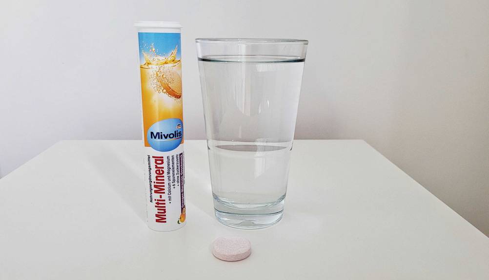 Eine Röhrendose Mineral-Brausetabletten steht neben einem Glas Wasser, vor dem eine einzelne Tablette liegt.