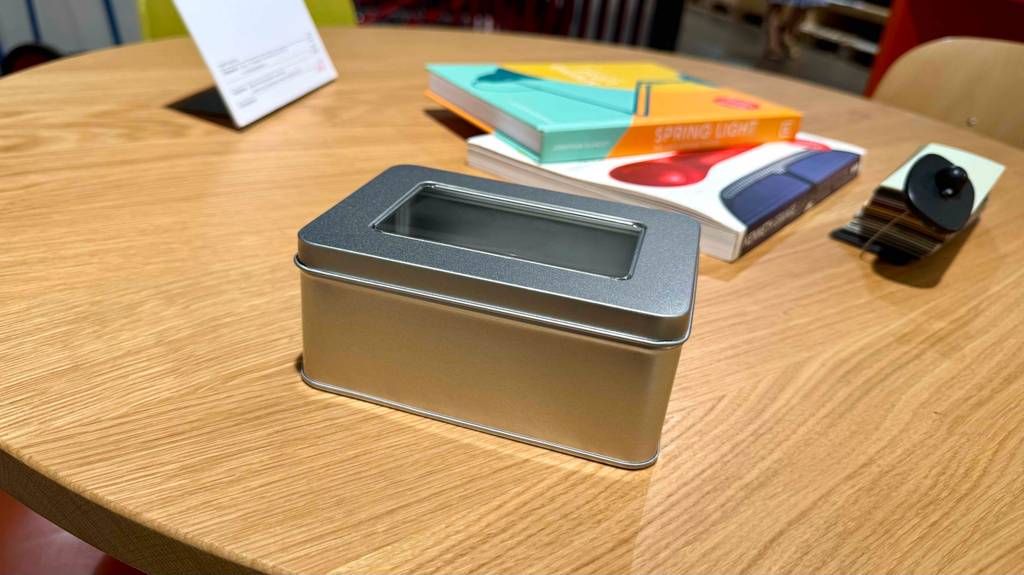 Die silberne, getestete Blechdose inklusive Deckel mit Sichtfenster steht auf einem Holztisch, auf dem zwei Bücher liegen.