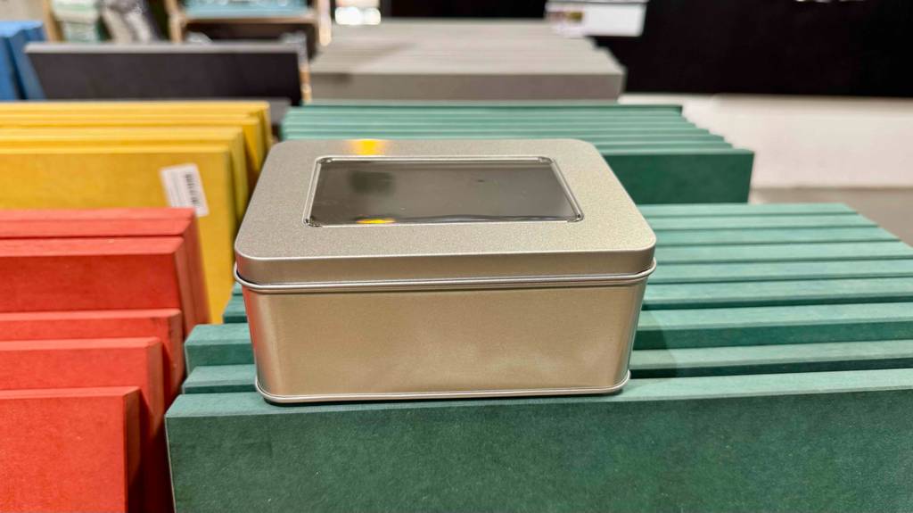 Im Test: Die Blechdose in silberner Farbe inklusive Deckel mit Sichtfenster steht auf mehreren grünen Spanplatten im Baumarkt.