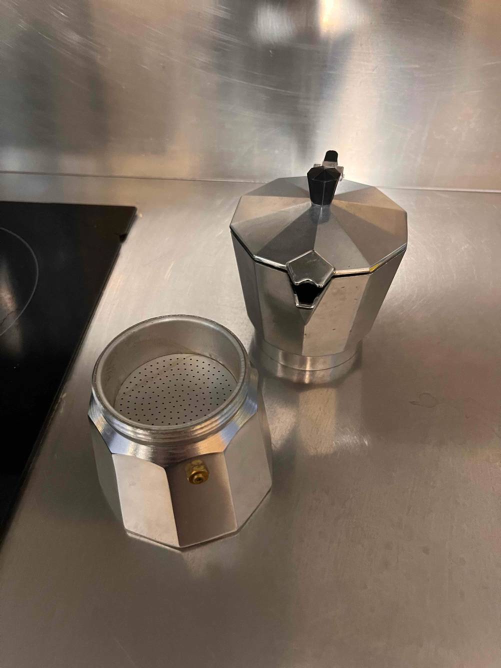 Metallfarbene, geöffnete Mokka-Kaffeemaschine im Test steht auf einer Aluminiumarbeitsfläche neben einem schwarzen Kochfeld.