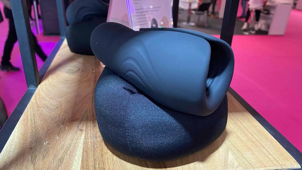Masturbator getestet: Schwarzer Masturbator Shooter lehnt auf einem schwarzen Ausstellungskissen in einem Holzregal, das auf einem rosa Boden steht.