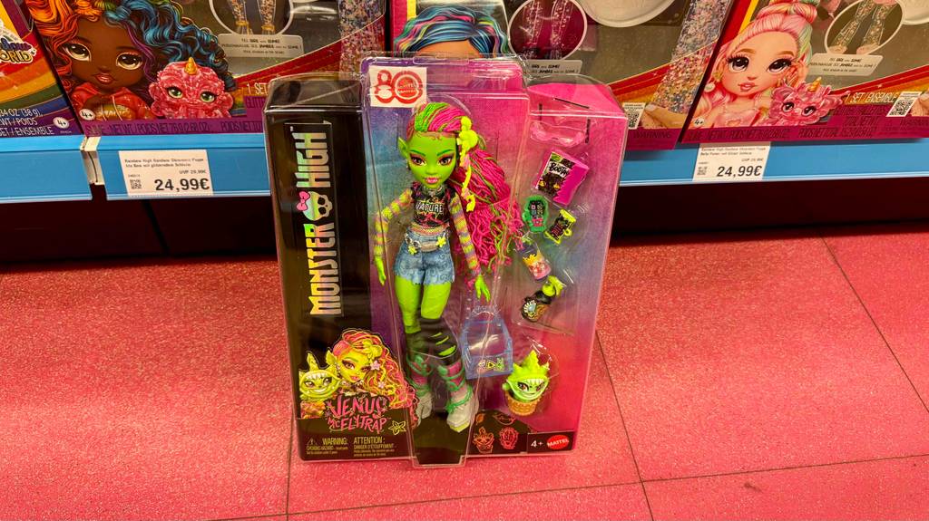 Eine getestete Monster-High-Puppe steht in Originalverpackung auf einem roten Fliesenboden vor einem Spielwarenregal.
