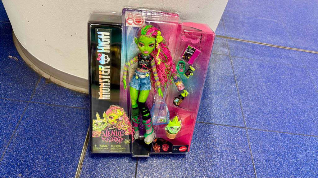 Monster-High-Puppen getestet: Eine Packung des Models Venus steht auf einem blauen Fliesenboden an eine Wand lehnend.